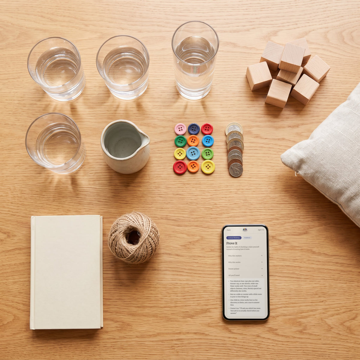 Everyday things for the activities laid out on a table: glasses of water, a jug, buttons, coins, wooden blocks, twine, with the Prove It activity on a phone